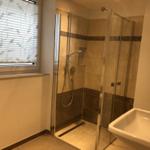 Badezimmer mit Dusche, Waschbecken und Fenster mit Jalousie.