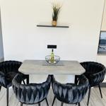 Dining area with table and four chairs, wall shelf with plant, and kitchen area in background.