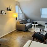 Wohnzimmer mit Sofa, Sessel und Holzboden. Fenster und Stehlampe sichtbar.