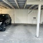 Black car parked under a carport with wooden frame and gray paving stones.