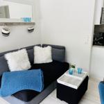 Schwarzes Sofa mit weißen Kissen und blauer Decke. Koffer-Tisch und Küchenbereich sichtbar.