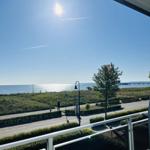 Balkon mit Blick auf Meer, Grünfläche und Promenade unter blauem Himmel.