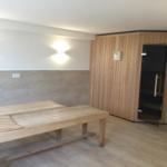 Sauna mit Holzbank und Tür. Wand mit Steinen und Licht.