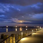 Lit wooden pier along the beach at night with moon and waves.