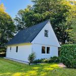 Weißes Haus mit dunklem Dach, umgeben von Gras und Bäumen.