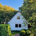 Weißes Haus mit Dachfenster, umgeben von Bäumen und Grünflächen.