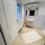 Badezimmer mit Waschbecken, Spiegel und Dusche. Fenster mit Vorhang und Teppich auf dem Boden.