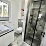 Badezimmer mit Dusche, Waschbecken und Toilette. Fenster mit Blick auf Bäume.