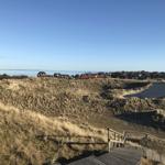 Holzsteg führt durch Dünen zu einem Stranddorf mit roten Häusern und Blick aufs Meer.