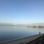 Ein Mensch in roter Jacke steht am Strand neben einem Steg und einem Hafen.