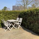 Holzterrasse mit Tisch und vier Stühlen, umgeben von grünen Hecken.