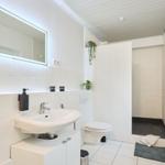 Badezimmer mit Waschbecken, Toilette und Dusche. Weiße Fliesen und beleuchtetes Spiegel.