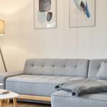 Ein grauer Sofa mit Kissen und Decke. Hinter dem Sofa hängen drei Bilder. Links steht eine Stehlampe und eine Pflanze.