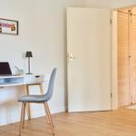 Arbeitsplatz mit Schreibtisch, Stuhl und Laptop. Tür führt in Schlafzimmer mit Holzschrank.