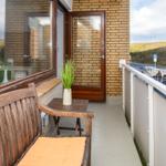Balcony with wooden bench, table, and plant. View of street and hills.