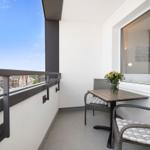 Balkon mit Tisch, zwei Stühlen und Blumenarrangement. Blick auf Wohngebiet unter blauem Himmel.