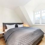 Schlafzimmer mit Dachschrägen, Bett mit grauer Bettdecke und Fenster mit Jalousien.