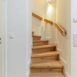 Holzgetreppe mit Geländer führt nach oben. Weiße Wände und Lichtschalter sind sichtbar.