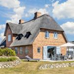 Ein Haus mit Strohdach, Terrasse und Garten. Ein Liegestuhl und Tisch sind draußen.