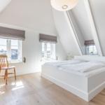 Schlafzimmer mit Dachschräge, weißem Bett, Holzboden und zwei Fenstern mit Jalousien.