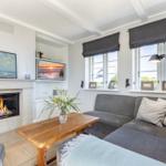 Wohnzimmer mit Kamin, Sofa und Fenstern mit Blick nach draußen.