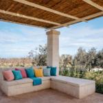 Terrace with stone bench, colorful cushions, and thatch roof. View of olive groves.