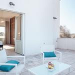 Terrasse mit weißen Stühlen und Tisch. Blick in Schlafzimmer mit Bett und Vorhängen.