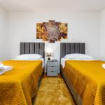 Zwei Betten mit gelben Bezügen und grauen Kopfenden. Zwischen ihnen steht ein Nachttisch mit Lampe.