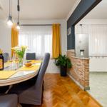 Essbereich mit runder Tisch, gelben Tischdecken und grauen Stühlen. Hintergrund: Küche mit Kühlschrank und Steinwand.