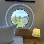 Schlafzimmer mit runden Fenster und Holzbett. Lampe und Blick auf Außenbereich.