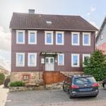 Zweistöckiges Haus mit braunem Holzverkleidung und weiß gerahmten Fenstern. Vorderhof mit Parkplätzen.