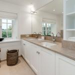 Badezimmer mit zwei Waschbecken, Spiegeln und Fenster. Holzstuhl und Korb sind vorhanden.