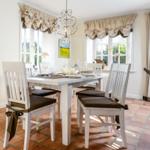 Esszimmer mit weißem Tisch, Stühlen und Terrakottboden. Fenster mit Vorhängen und Kronleuchter.