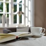 Ein Buch und eine Tasse stehen auf einem Tisch vor einem Fenster.