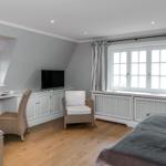 Schlafzimmer mit Bett, Schreibtisch, Sessel und TV. Holzboden und große Fenster.