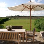 Terrasse mit Tisch, Stühlen und Sonnenschirm. Grünfläche und Felder im Hintergrund.