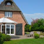 Rotes Backsteingebäude mit Strohdach, Terrasse mit Sonnenschirm und Liegestühlen im Garten.