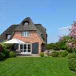 Haus mit Strohdach, Garten, Sonnenliegen und blühender Baum.