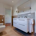 Badezimmer mit Waschtisch, Spiegel und Toilette. Warme Fliesen und weißer Schrank.