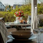 Terrasse mit Esstisch, Blumen und Frühstück. Blick auf grüne Hecke und Häuser.