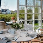 Runder Tisch mit Geschirr und Gläsern vor großem Fenster mit Blick auf Garten.