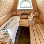 Sauna mit Holzbank, Steinmulde und Eimer. Fenster mit Bogenform. Decke und Wände aus Holz.