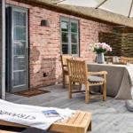 Terrasse mit Holzgartenmöbeln, Tisch mit Blumen und Sonnenschirm. Steinmauer und Ziegelwand im Hintergrund.