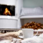 Eine weiße Tasse steht auf einem Fell, vor einem Kamin mit brennendem Feuer.