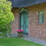 Ein roter Ziegelbau mit grüner Tür und Dach aus Stroh. Vor dem Eingang steht ein Blumentopf.