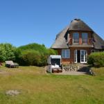 Rotes Backsteingebäude mit Strohdach, Terrasse und Garten mit Liegen.