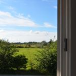 Durch das Fenster blickt man auf eine grüne Wiese und einen blauen Himmel mit weißen Wolken.