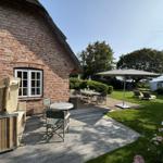 Holzterrasse mit Tischen, Stühlen und Sonnenschirm neben einem Backsteinhaus mit Dach.