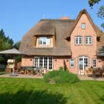 Haus mit Strohdach, Terrasse und Garten mit Liegestühlen und Tischen.