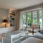 Wohnzimmer mit Sofa, Sessel, Fernseher und Bücherregal. Große Fenster mit Blick auf Garten.